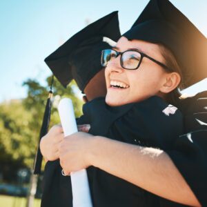 Wearing caps and gowns, two graduates embrace outdoors as one holds a Ut Sed Elementu diploma, sunlight streaming in the background.