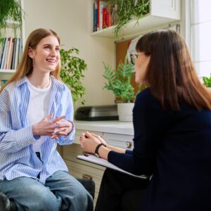 Two people are having a conversation in a bright room filled with books and plants, with one holding a Lorem Ipsum Dolor notebook.
